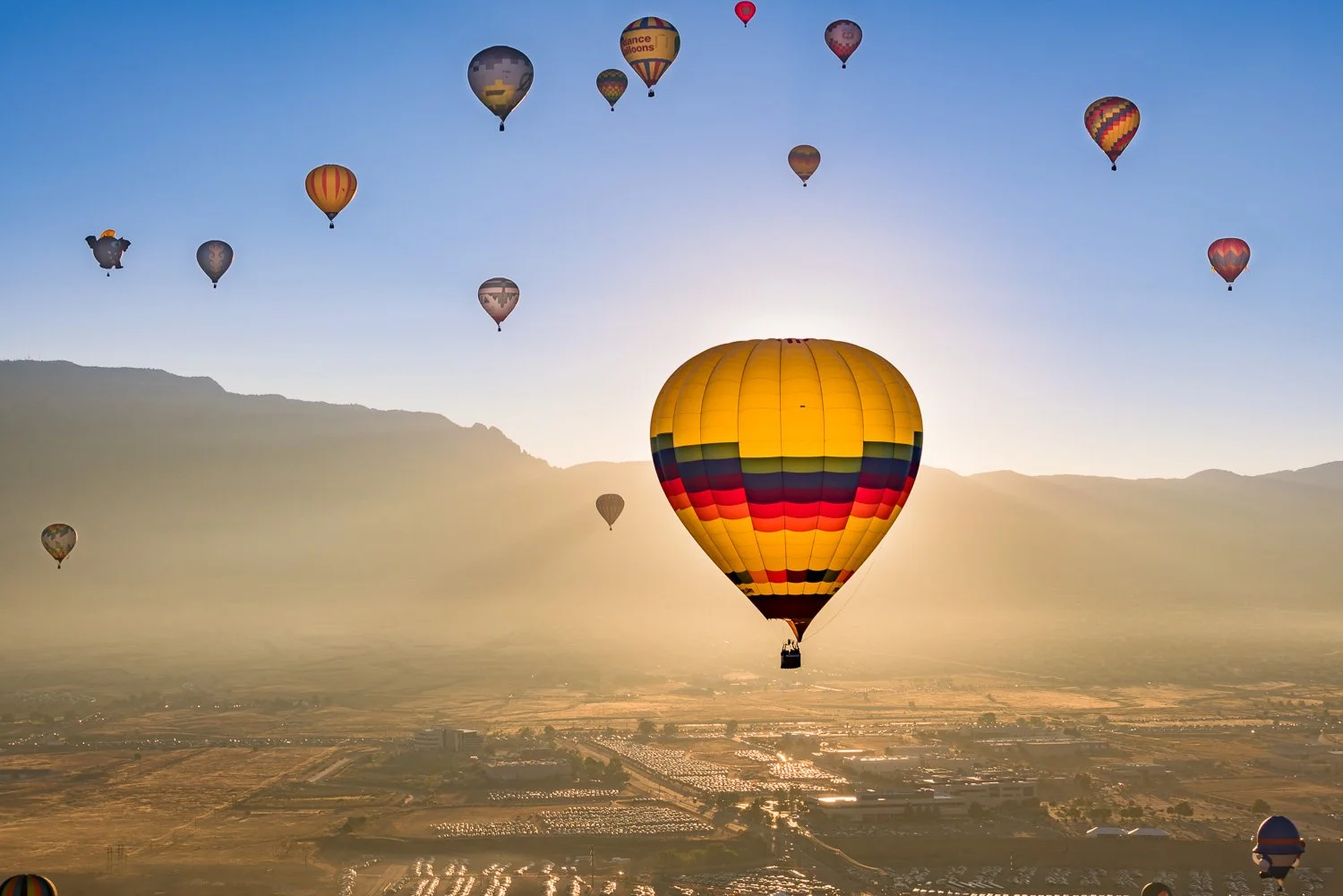 Several colorful hot air balloons floating in the sky during sunrise or sunset over a valley with mountains in the background.