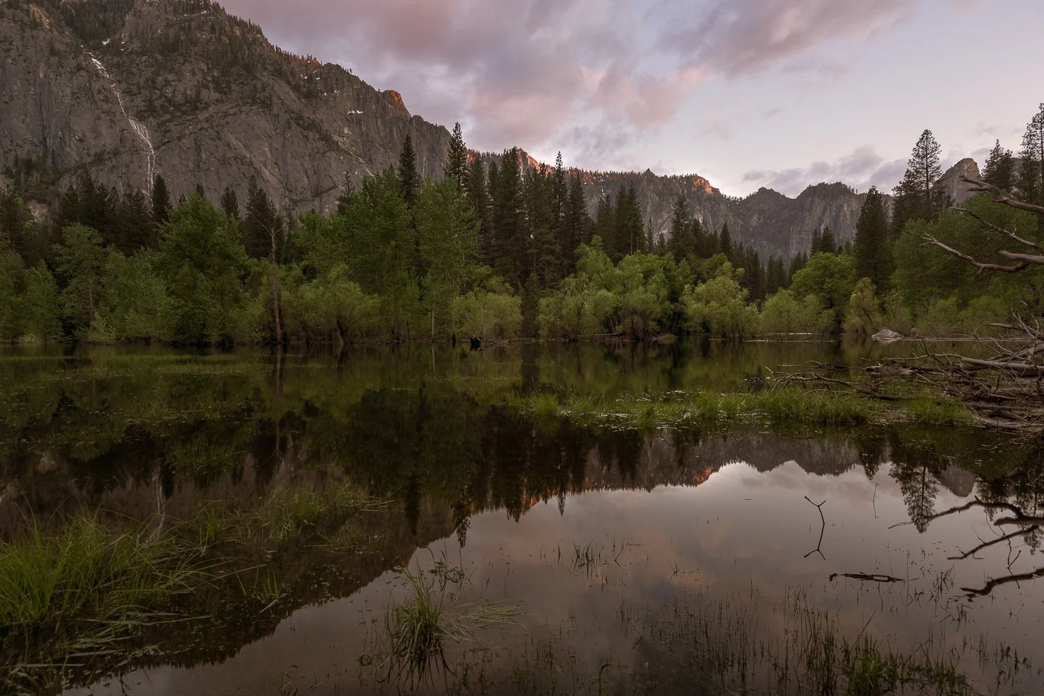 A tranquil landscape with a calm river reflecting the surrounding green trees and mountains during sunset.