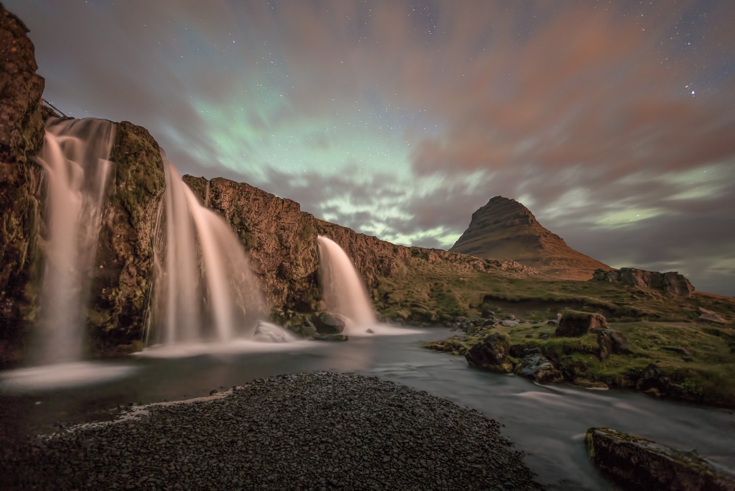Nighttime landscape featuring Kirkjufell mountain, waterfalls, and the Northern Lights in Iceland.