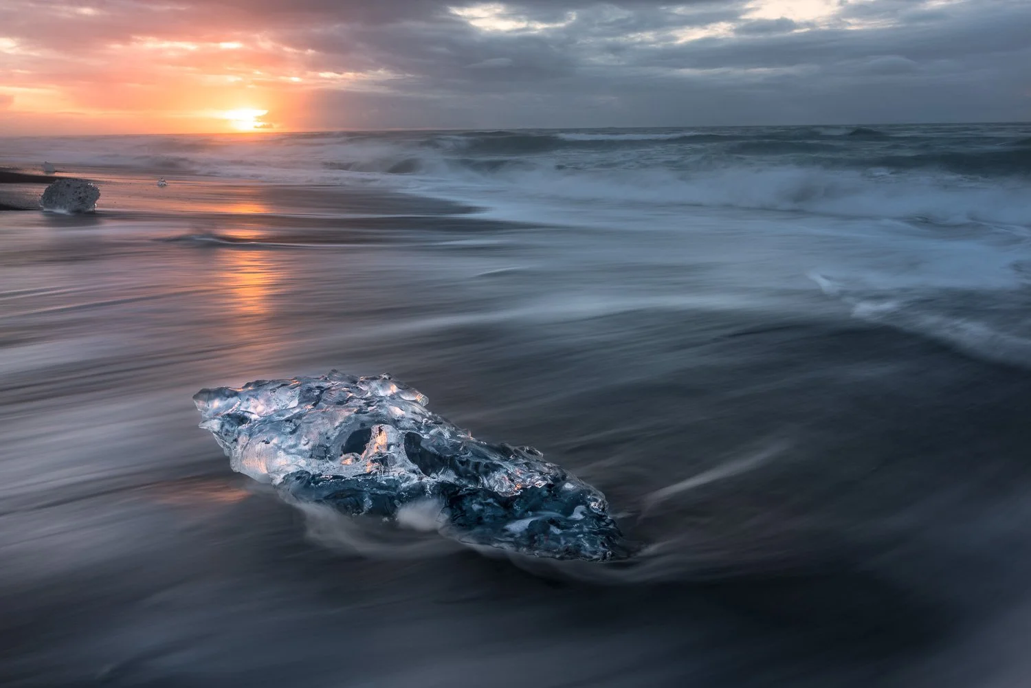 A large piece of ice on a beach at sunset, with waves and a cloudy sky in the background.