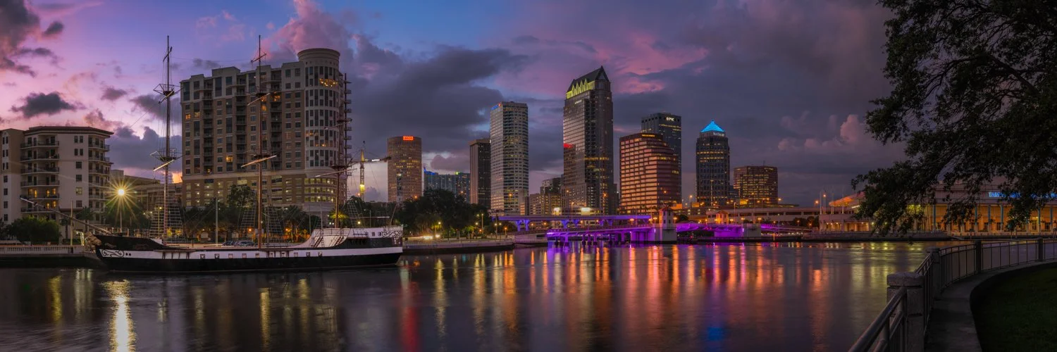 City skyline at dusk with high-rise buildings, a historic tall ship docked on the river, and colorful reflections on the water.