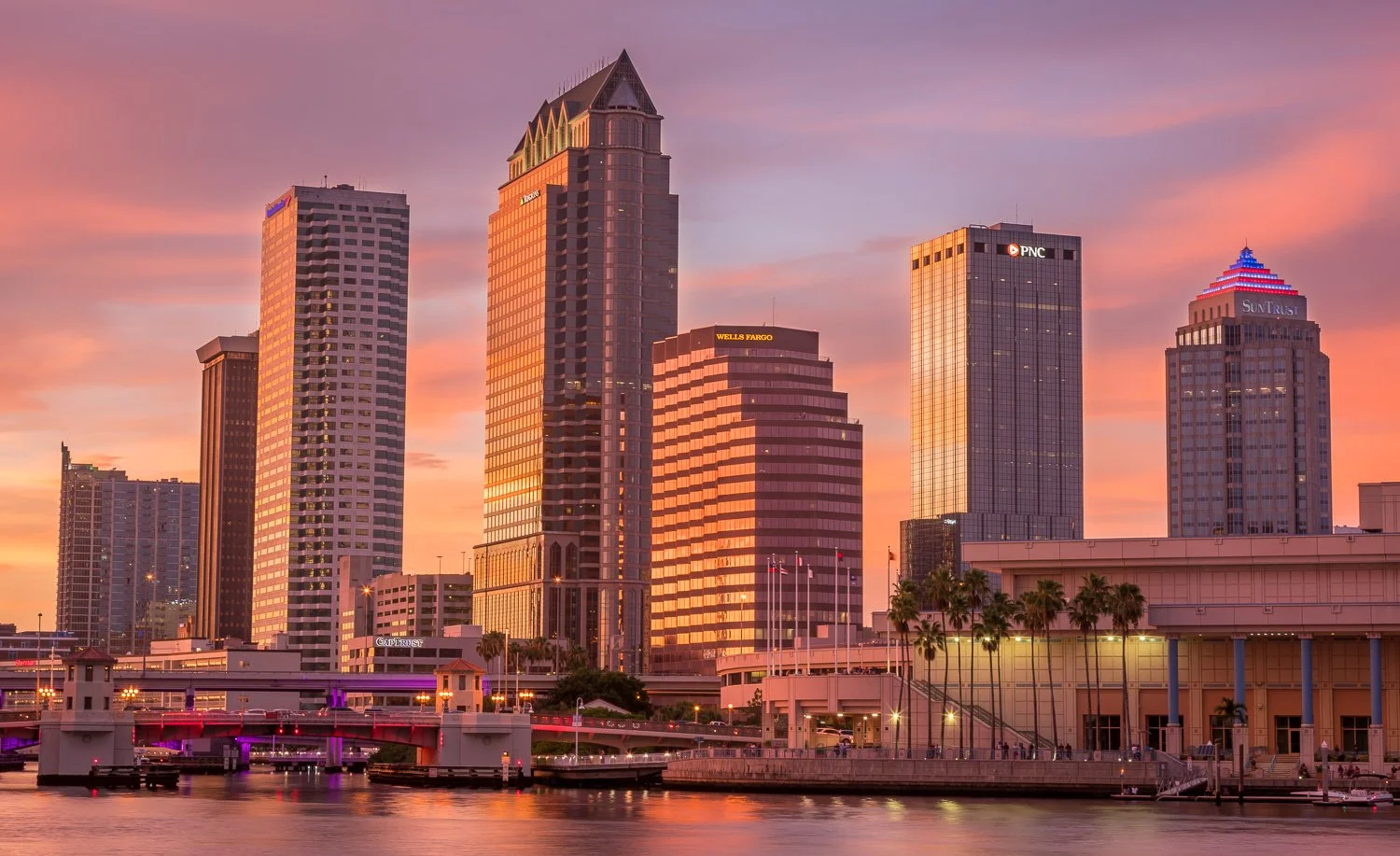 Sunset skyline of Tampa, Florida with tall modern skyscrapers reflecting the pink and orange sky, palm trees, water, and a bridge in the foreground.