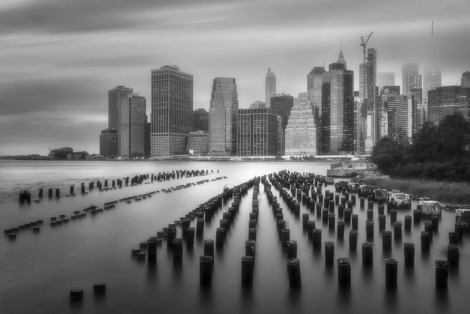 Black and white photo of Manhattan skyline with buildings under cloudy sky, seen across a body of water with weathered wooden posts and remnants of pilings in the foreground.