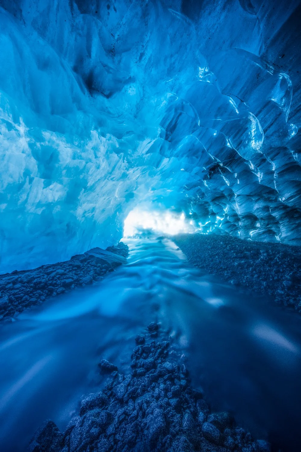Image of the interior of a blue ice cave with water flowing through rocky floor.