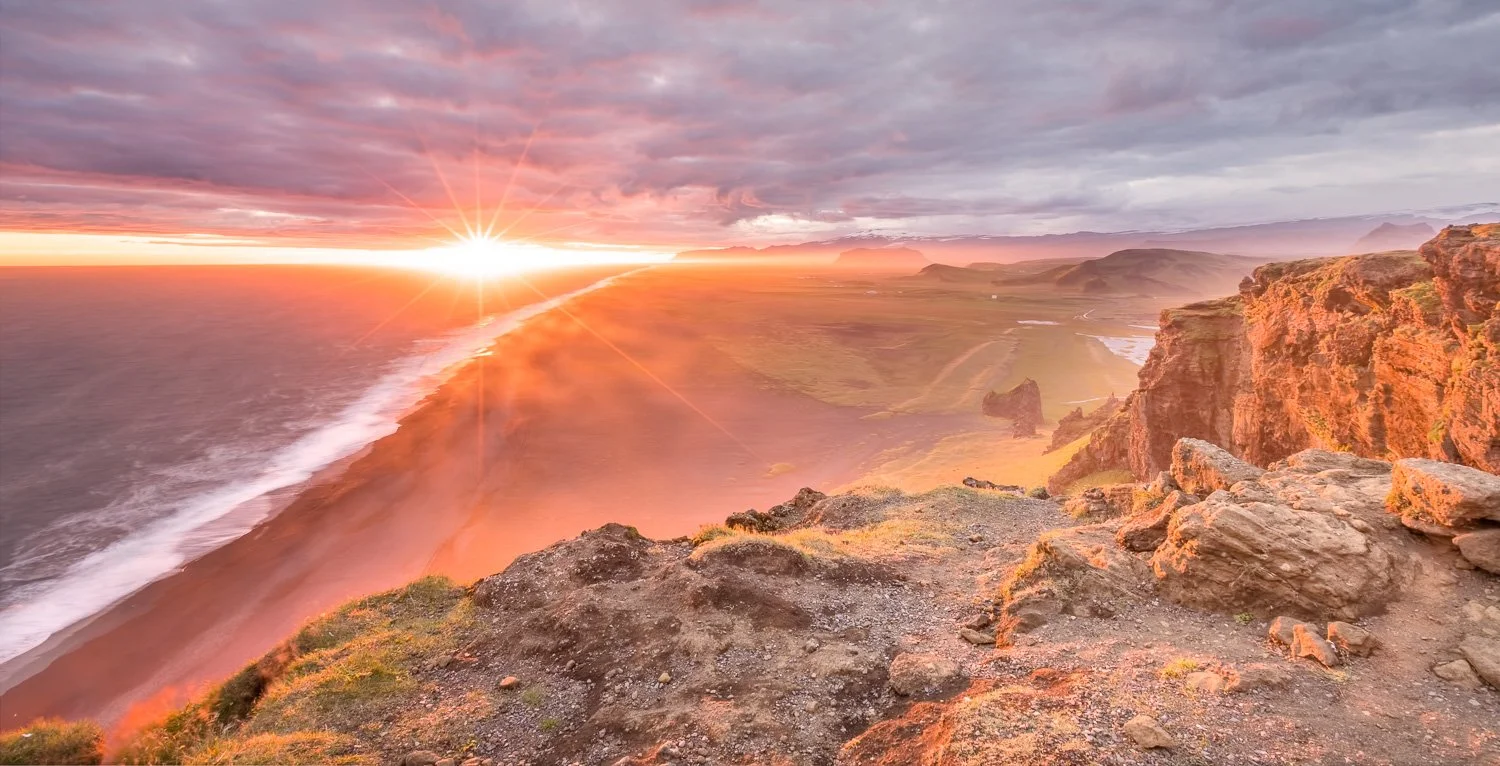 Sunset over rugged coastline with cliffs and ocean waves, with clouds and distant mountains in the background.  This is from Iceland
