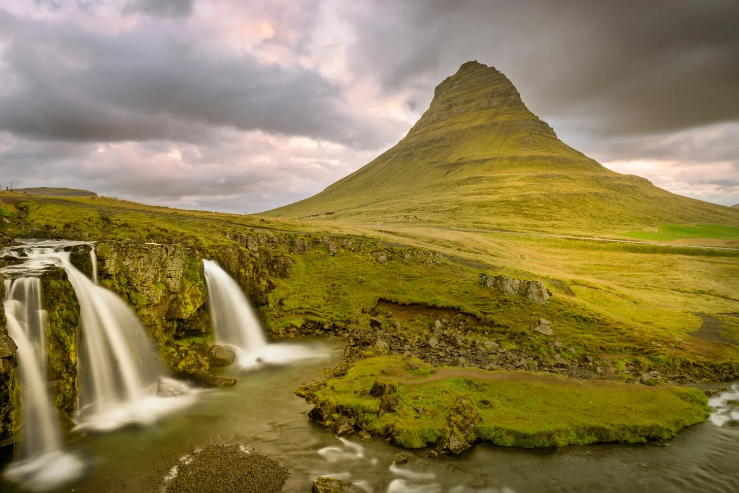A scenic landscape featuring the famous Iceland conical mountain, Mount Kirkjufel, a waterfall cascading over rocks, green grassy land, and a cloudy sky.