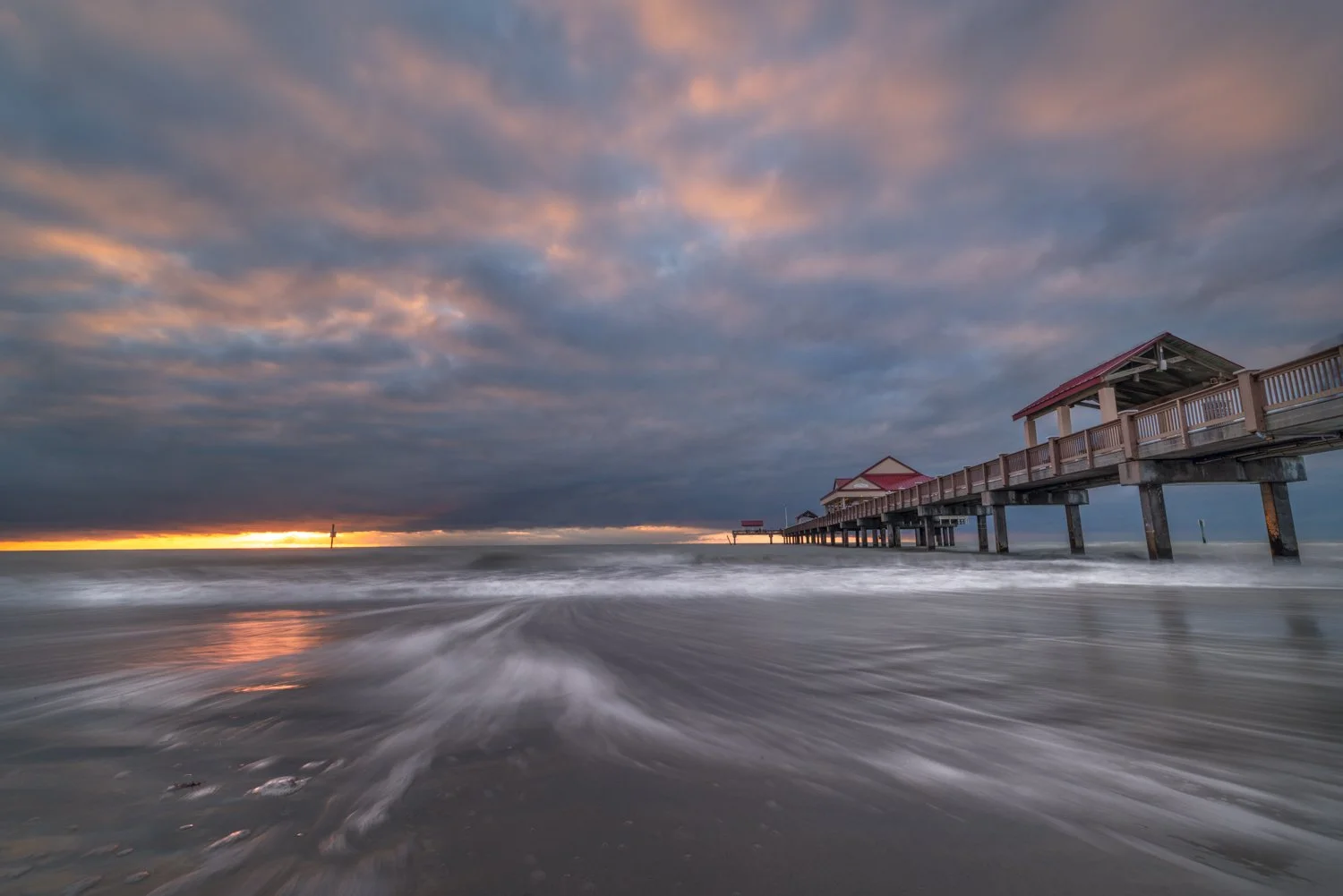 Sunset over the ocean with a cloudy sky.  This is Pier 60 in Clearwater Beach, Florida where the pier extends into the water, and waves crashing on the shore.