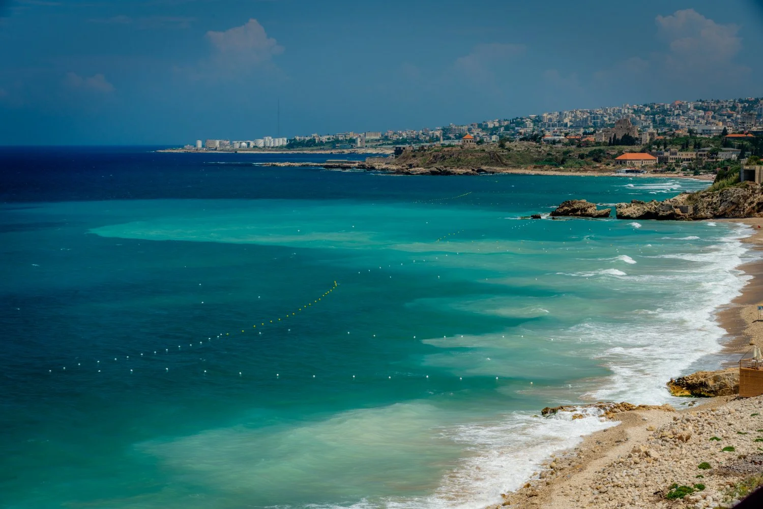 A coastal scene with turquoise waters, rocky shoreline, and a distant cityscape under a partly cloudy sky.  The village is the ancient port town of Byblos on the Mediterranean Sea in Lebanon.  The Greeks associated the city with the export of papyrus