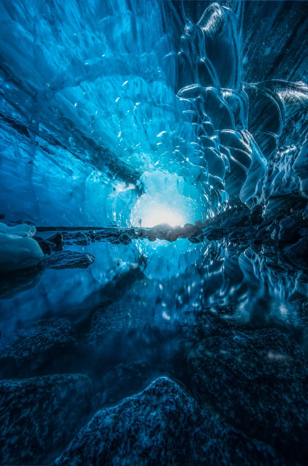Inside an icy glacier cave with blue ice formations and a small pool of water reflecting the ice, with light coming from the cave's opening.