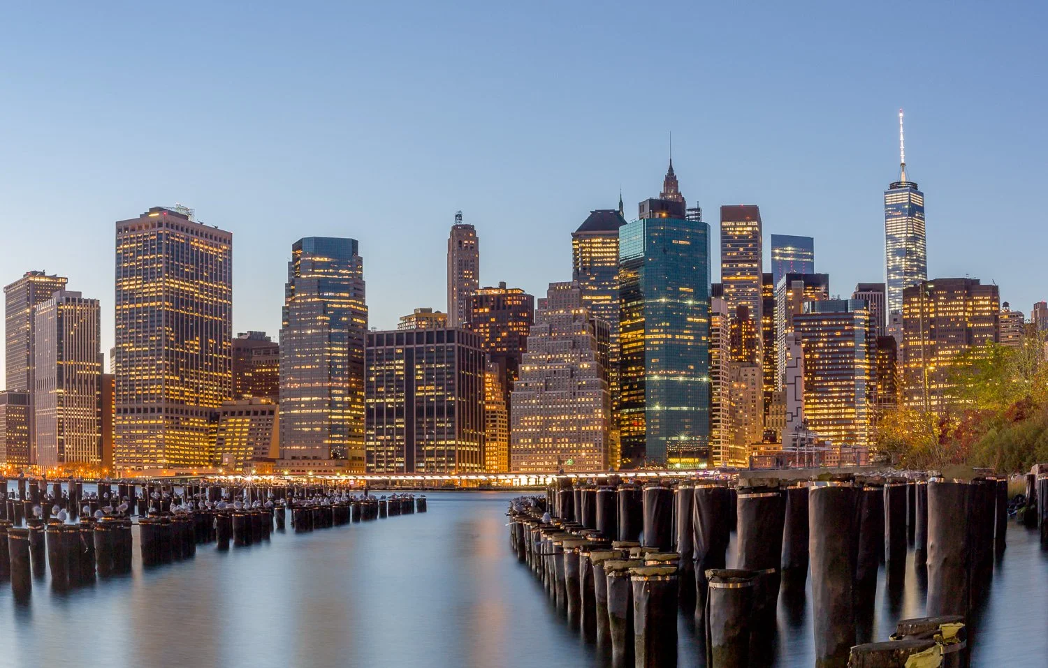 Lights illuminate skyscrapers in Manhattan skyline at dusk, viewed from waterfront with wooden pilings in the foreground.