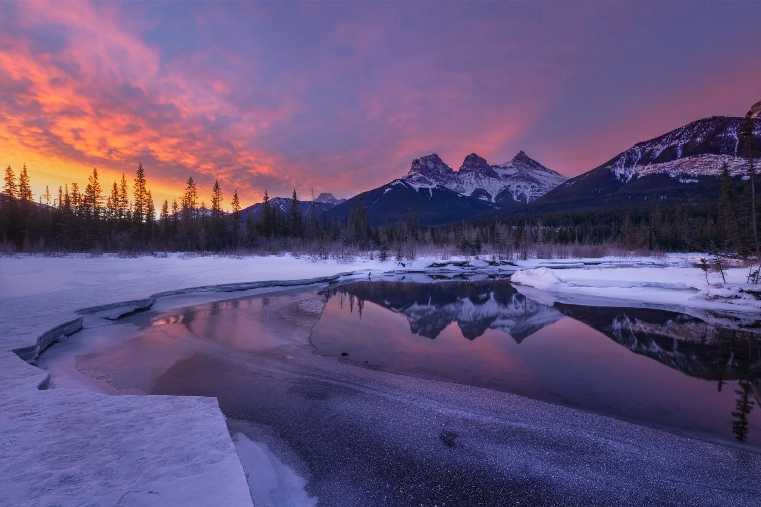 Snow-covered landscape with a partially frozen river reflecting a colorful sunset sky, surrounded by pine trees and mountains in the background.