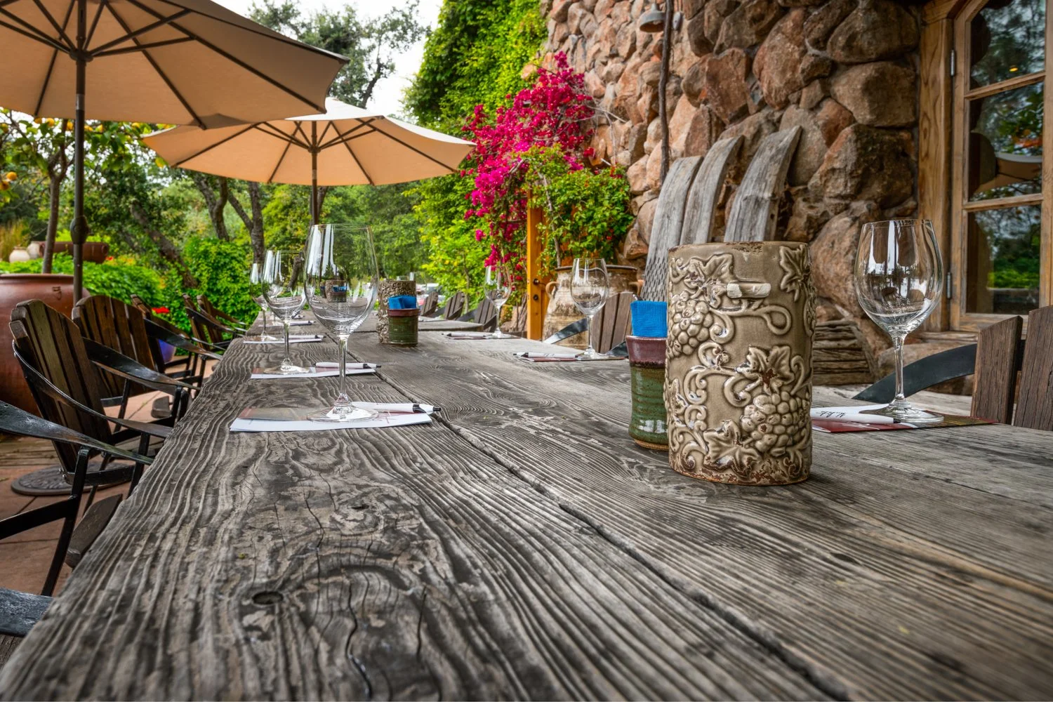 Outdoor dining table set with wine glasses, napkins, and ceramic vases, surrounded by chairs and umbrellas, with greenery, flowering plants, and a stone wall in the background. This shot was taken at Culeto Winery in Napa Valley