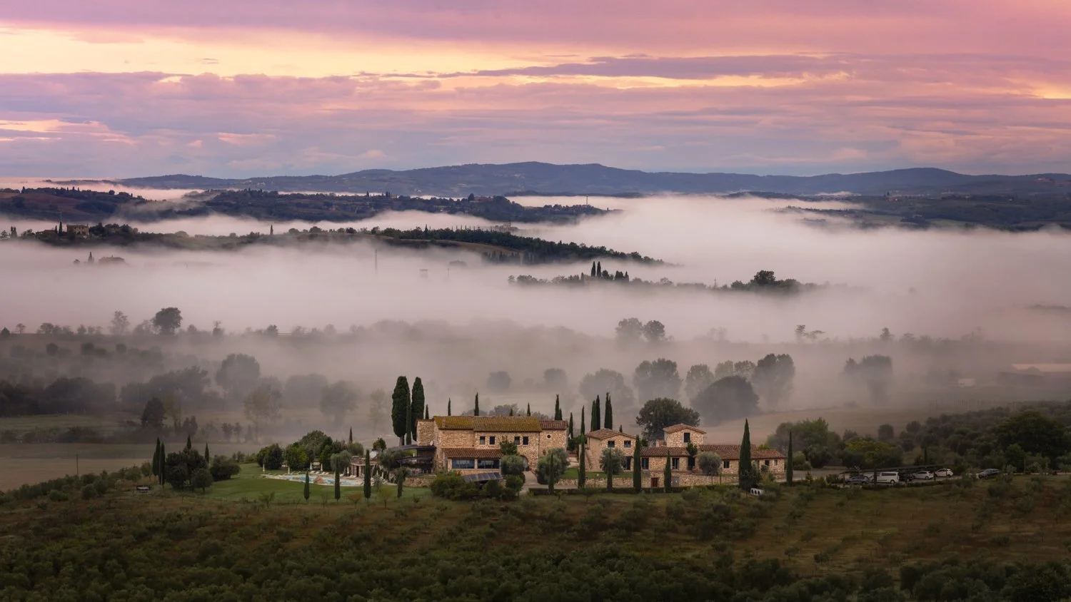 A scenic landscape of a countryside estate, a Tuscan field with stone buildings surrounded by cypress trees, set against rolling hills and mist, during dawn with pink and purple sky.  I took this shot from an overlook at the edge of Serra Del Terre, 
