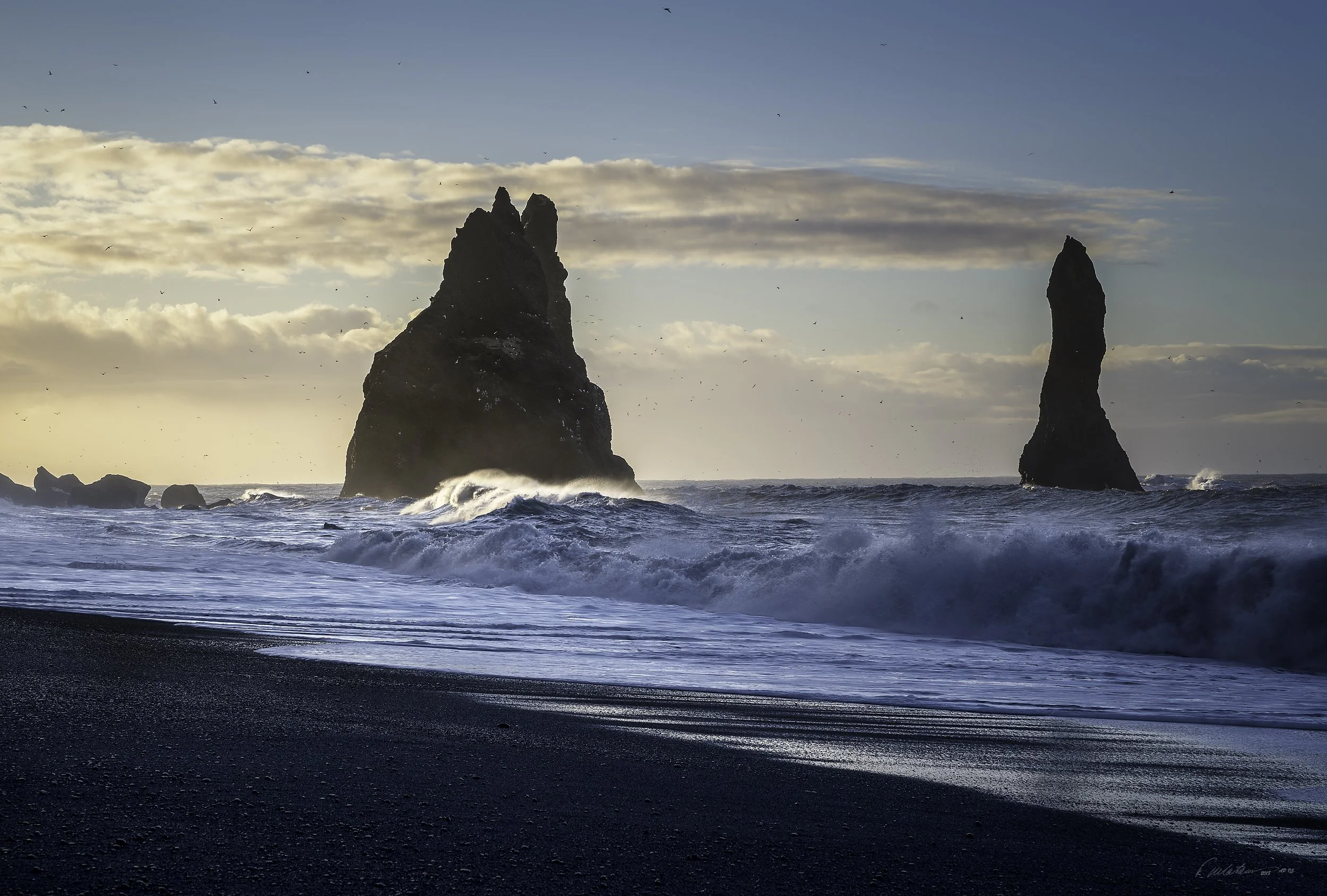 Ocean waves crashing against a black pebble beach with two large sea stacks in the background, under a partly cloudy sky