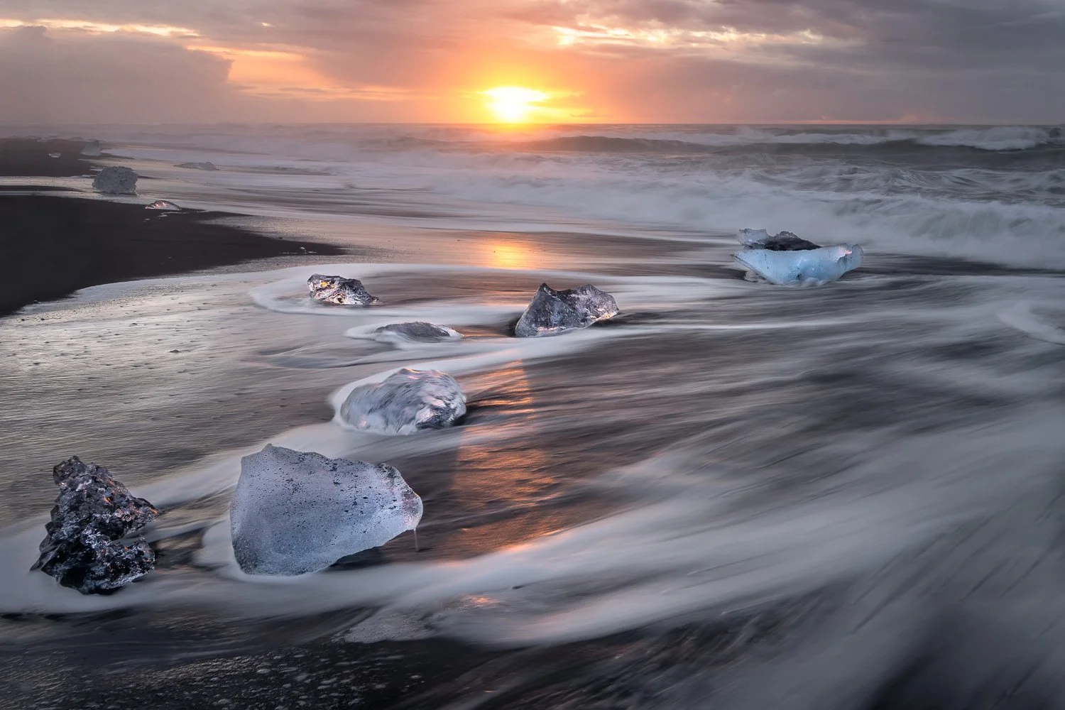 Sunset over a black sand beach with scattered ice rocks and waves crashing, creating a tranquil and cold landscape.