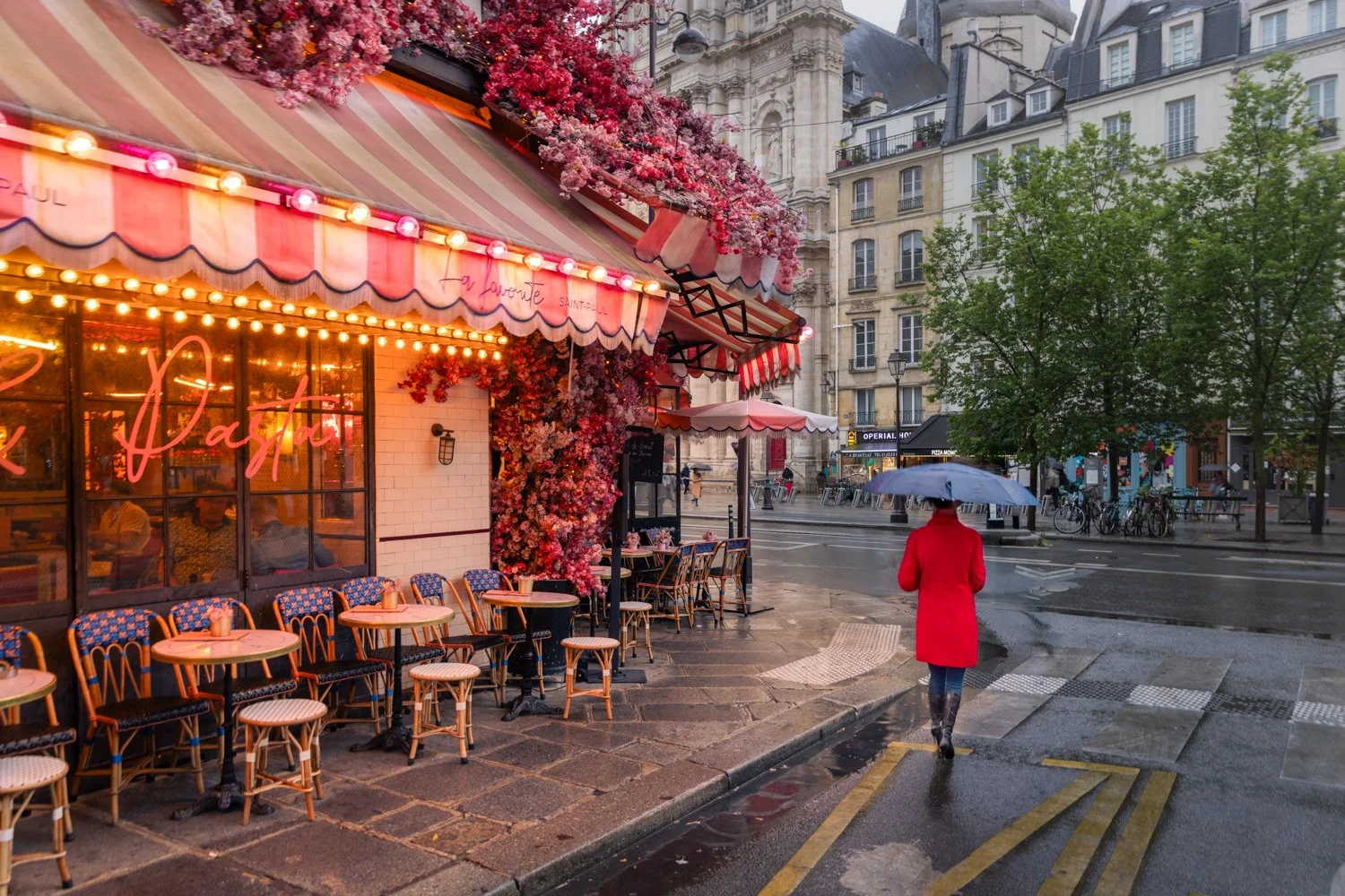 A woman in a red coat and black boots walking with an umbrella on a rainy street in front of a pink and red striped cafe with outdoor seating and pink flowers, in a rainy summer evening in Paris.