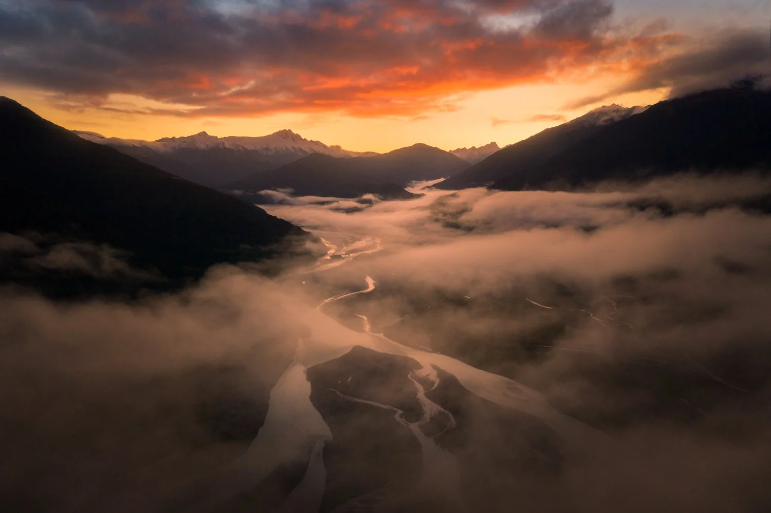 Aerial view of a river winding through a mountain valley at sunset, with fog over the river and snow-capped peaks in the distance under a colorful sky. Shot taken via drone photography in New Zealand