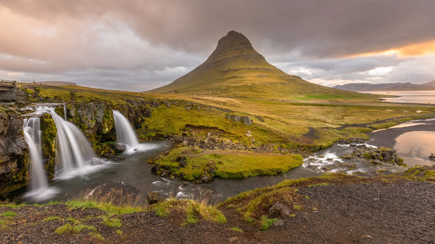 A scenic landscape featuring a tall, conical mountain, a small waterfall with cascading water, a river, and lush green grass under a cloudy sky during sunset.