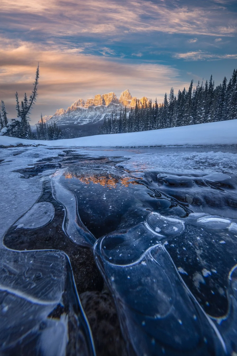 A winter landscape in the Canadian Rockies featuring a partially frozen river with ice formations, snow-covered banks, tall evergreen trees, and a mountain range illuminated by sunlight, with a colorful sky overhead.