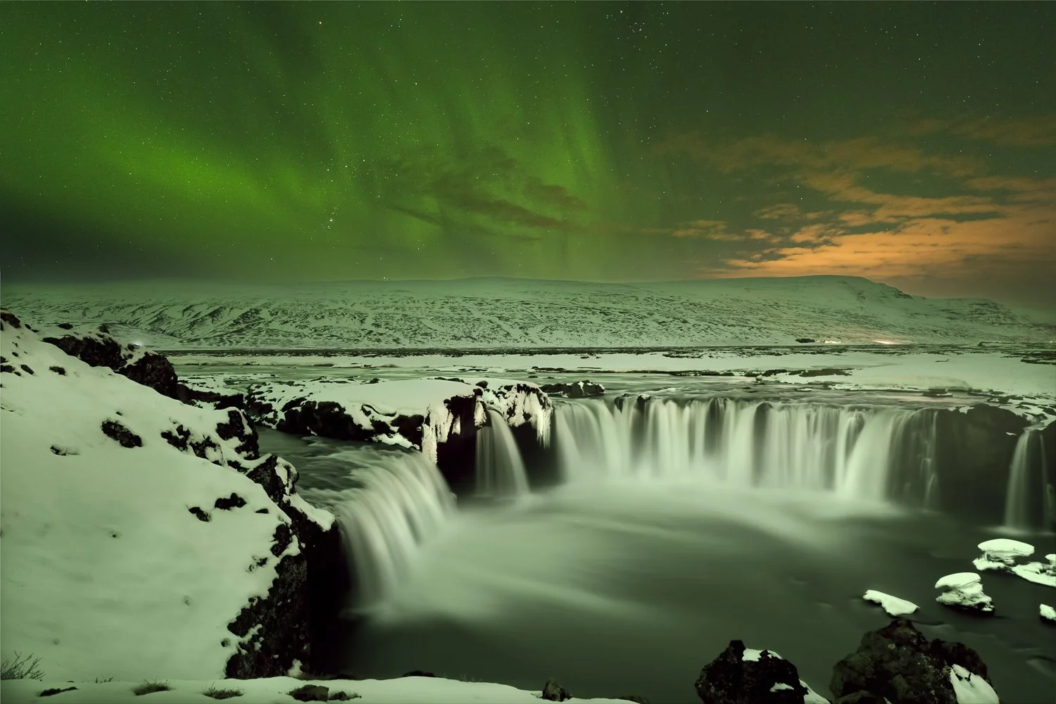Aurora borealis over a snowy landscape with a waterfall in Iceland at night.  The waterfall is called GodaFoss (Waterfall of the gods)