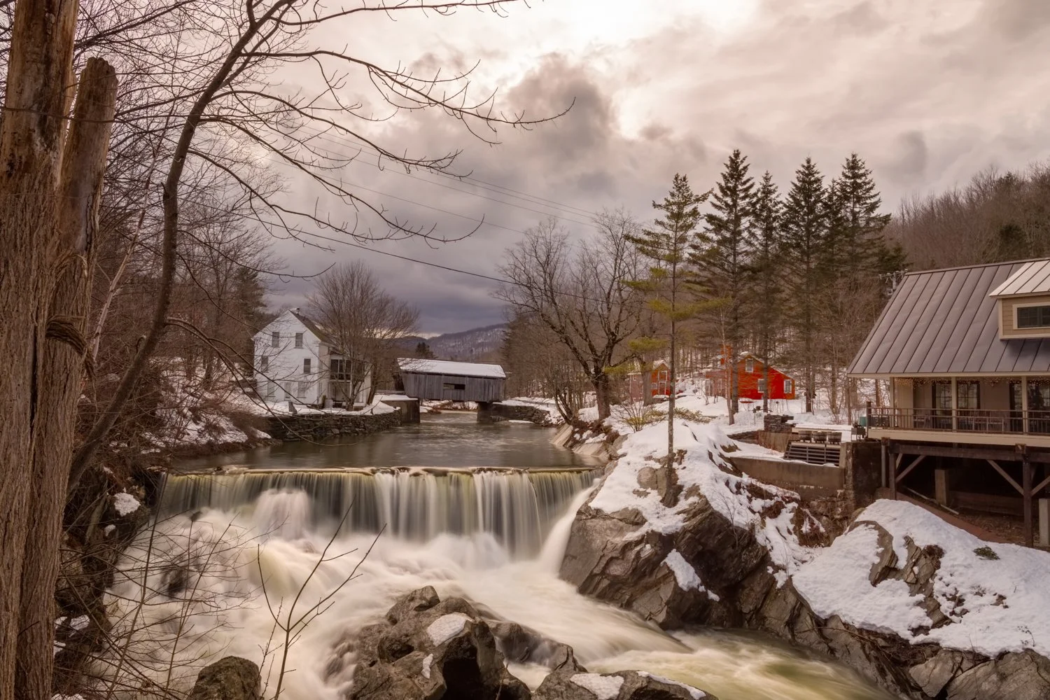 A winter scene in Vermont featuring a river with a small waterfall, snow-covered banks, and a tree in the foreground. In the background, there is a covered bridge (common in Vermont), a white house, a red barn, and a modern house with a metal roof. T