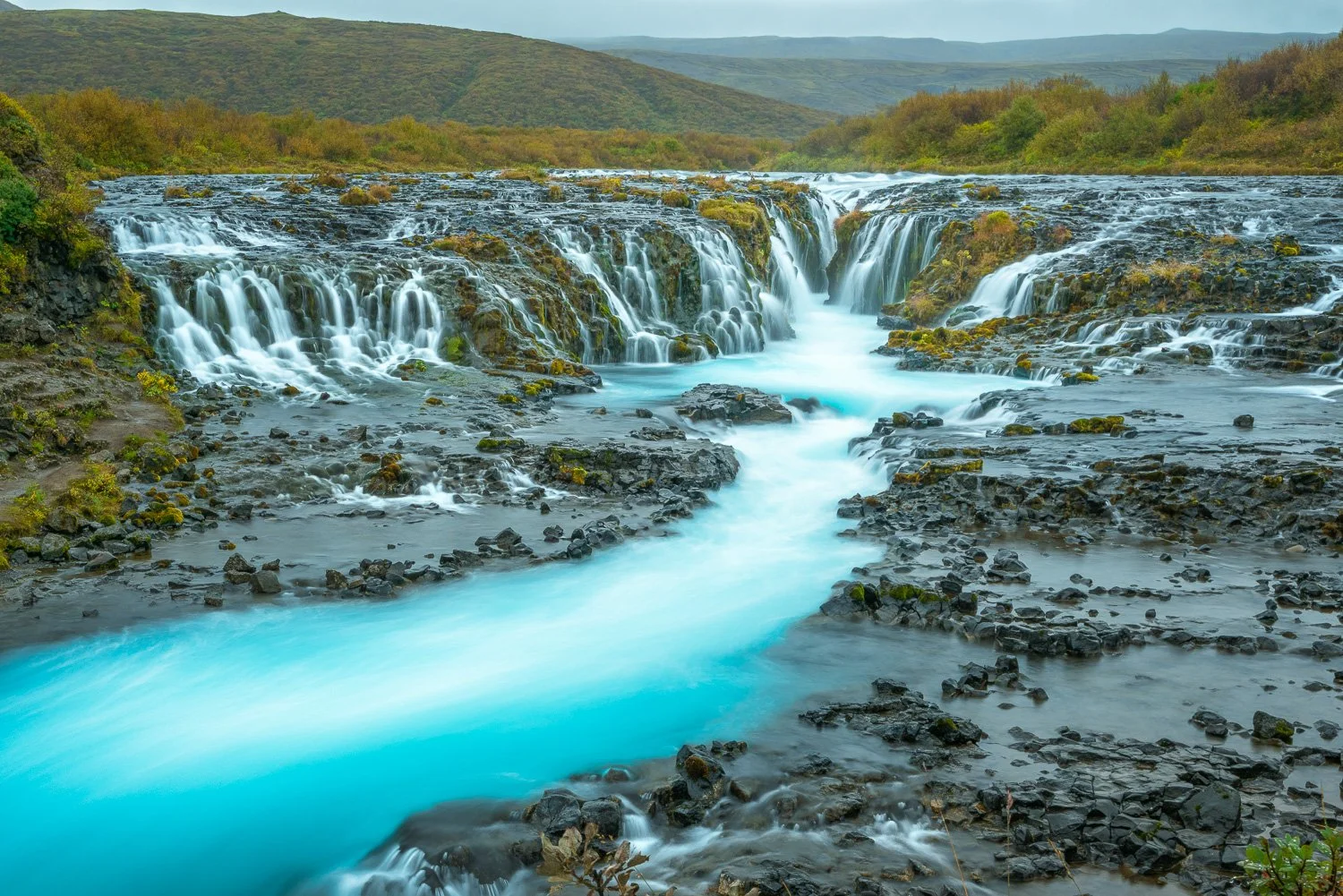 Waterfall cascading over rocky terrain with turquoise water flowing through a lush green landscape with hills in the background.