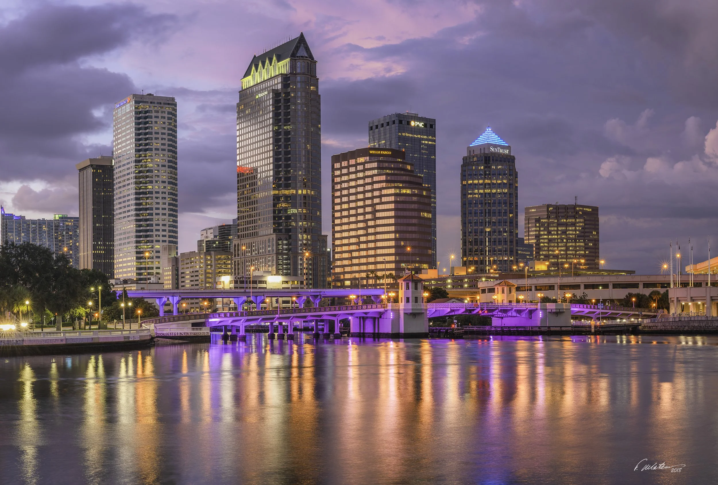 Sunset view of Tampa skyline with illuminated high-rise buildings and reflections on the water, featuring a bridge with purple lights in the foreground.
