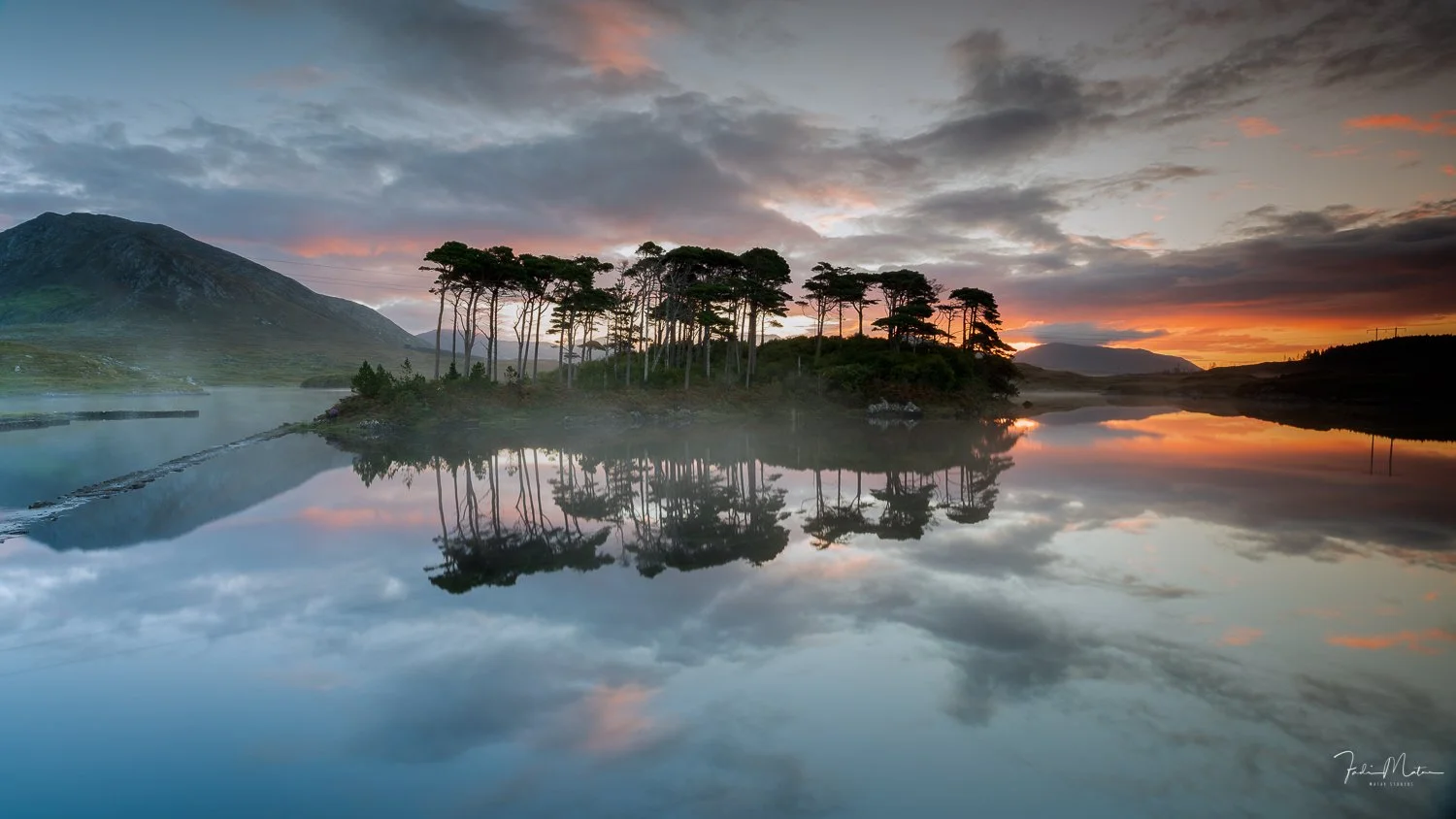 Scenic view of a small forested island in Ireland reflected in a calm lake at sunrise, with mountains and colorful sky in the background.