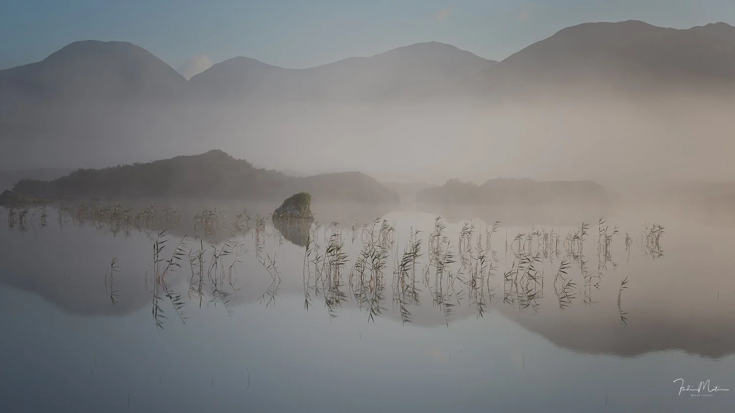 A misty lake with calm water reflecting the surrounding hills and mountains in the background. This is a calming sunrise that I witnessed in Ireland. There are reeds growing in the water and a large rock with greenery on it. The scene is tranquil and