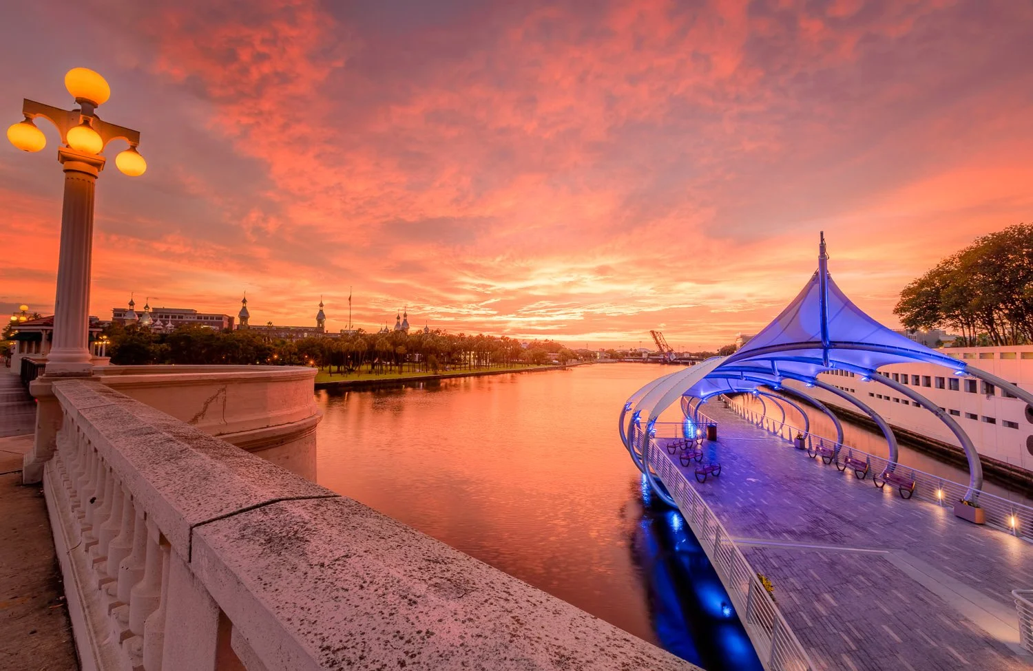 A scenic view of a river at sunset with a colorful sky, a white stone railing in the foreground, and a modern, illuminated blue pavilion structure on the dock extending over the water. This is River Walk in downtown Tampa