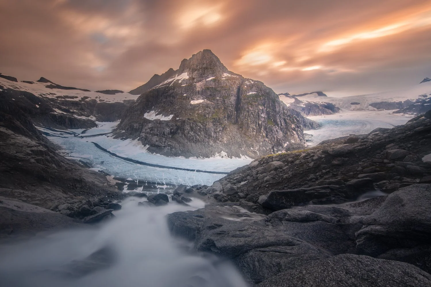 A rugged mountain landscape with snow patches, a glacier, and a flowing waterfall in the foreground, under a colorful cloudy sky in Alaska.  The waterfall is much steeper than it looks in this picture. 