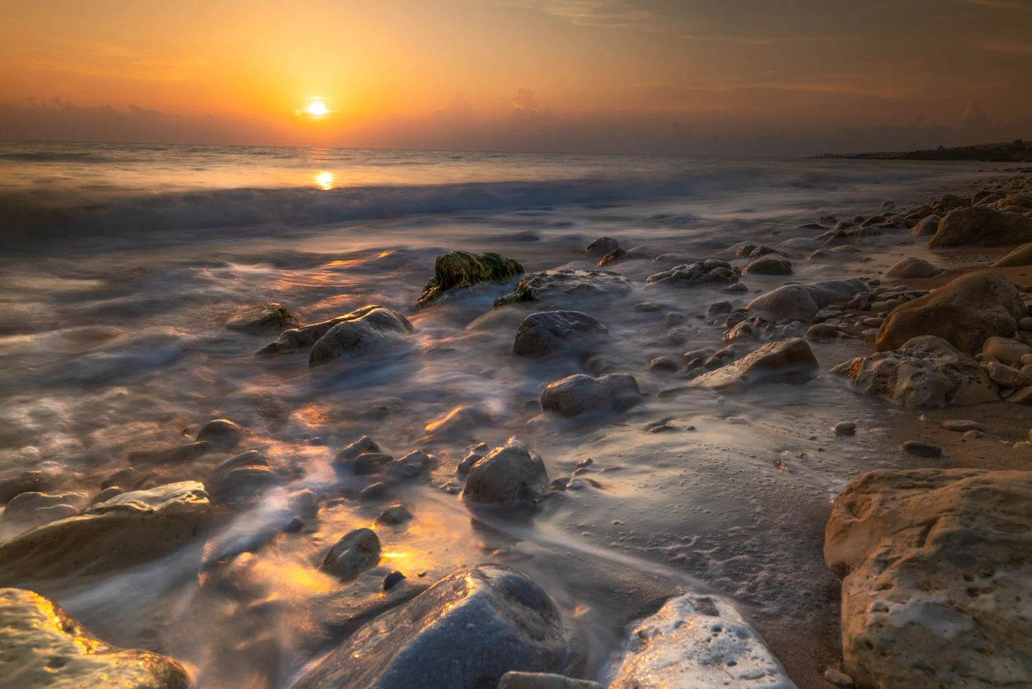Sunset over a rocky beach with calm waves washing over stones and sand on the Mediterranean sea in Lebanon.