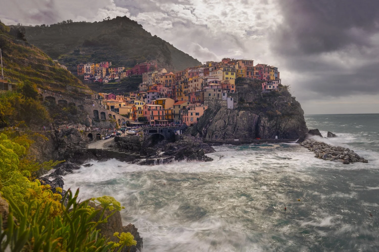 Colorful houses on a steep hillside in Cinque Terra overlooking the sea, with waves crashing against rocks and a cloudy sky.