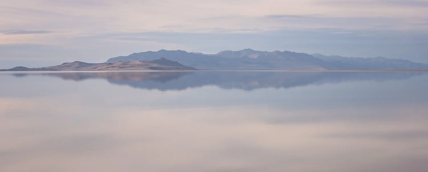 A calm body of water reflecting a mountain range under a partly cloudy sky.