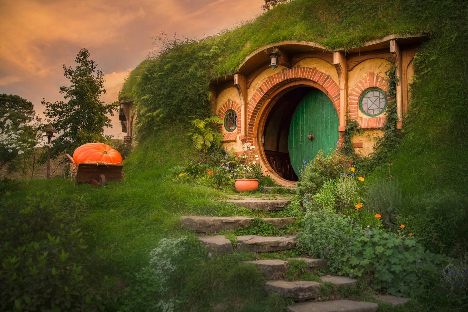 This is the cozy Bilbo Baggins hobbit house with a rounded green door and small circular windows, built into a grassy hillside. I took this shot at the Hobbiton movie set in New Zealand.  The stone steps lead up to the entrance, surrounded by colorfu