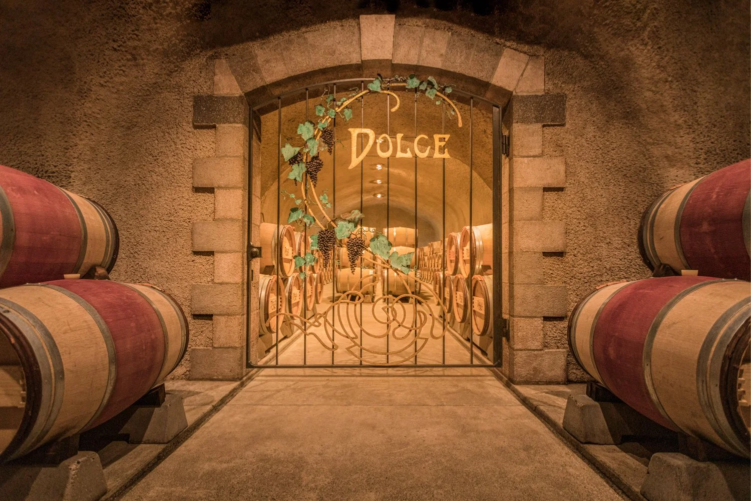 Wine cellar entrance with barrels stacked on either side, gated with a decorative iron gate featuring grapevine motif, and a sign reading 'DOLCE' above the gate.  This was taken in Far Ninete Winery