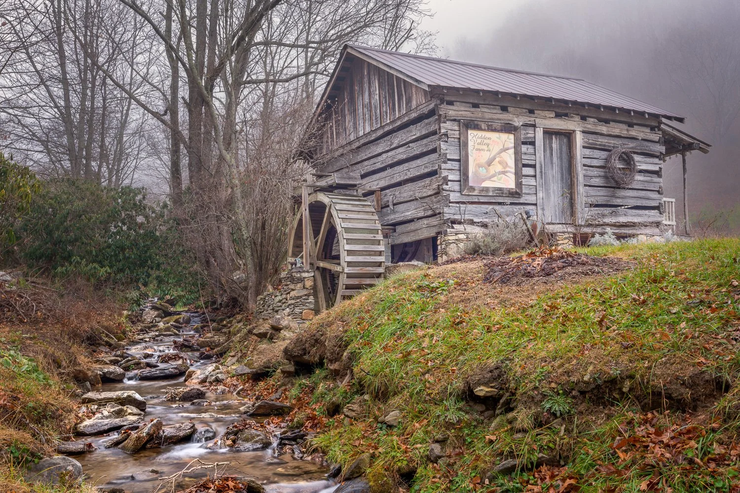 An old wooden watermill next to a small creek in a foggy, leafless forest.