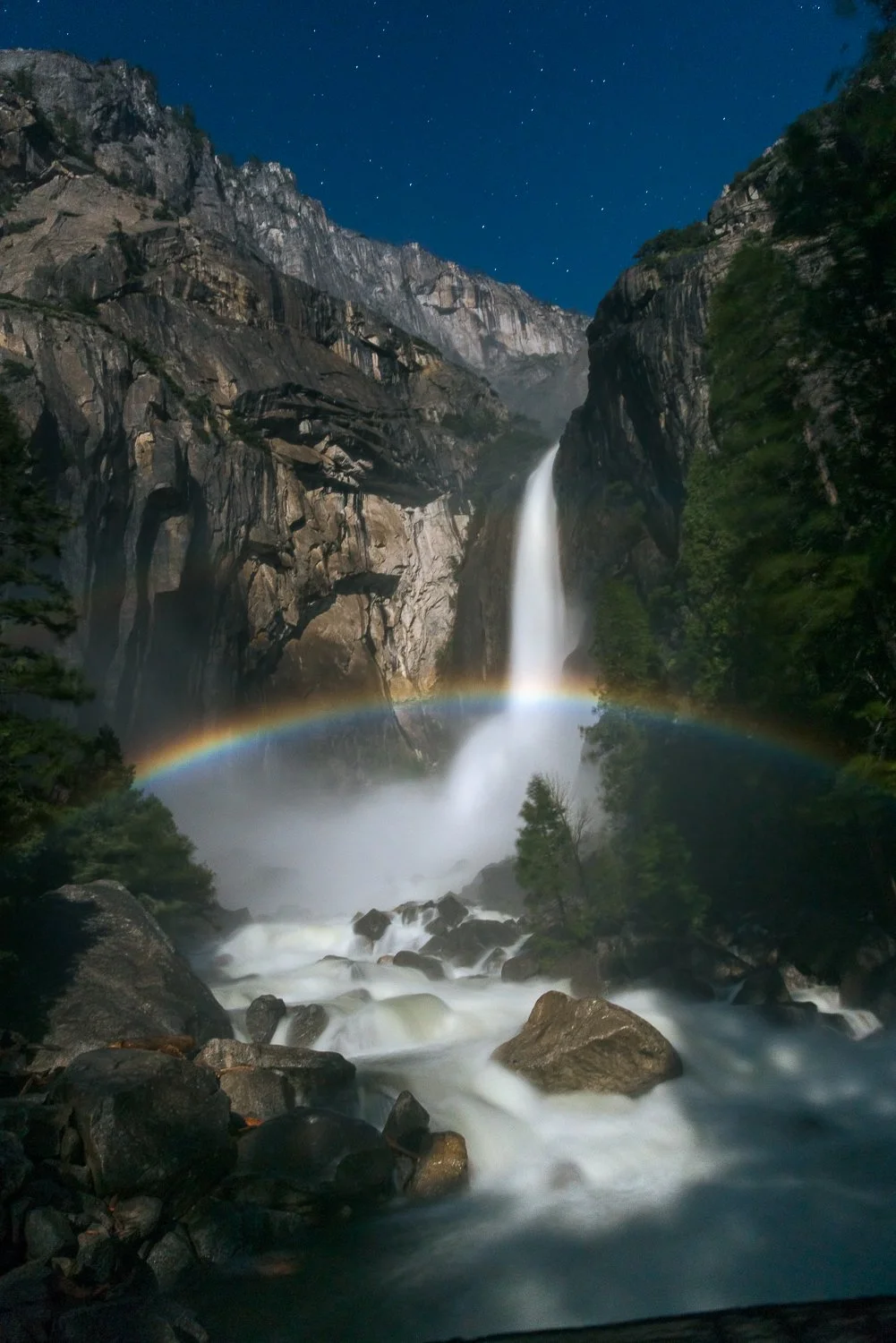 Nighttime photograph of a tall waterfall in a rocky canyon, with a rainbow forming at the base and a starry sky overhead.  This is in Yosemite and is called "Moon Bow". It happens close to midnight on a full moon day where the moonlight hits the mist