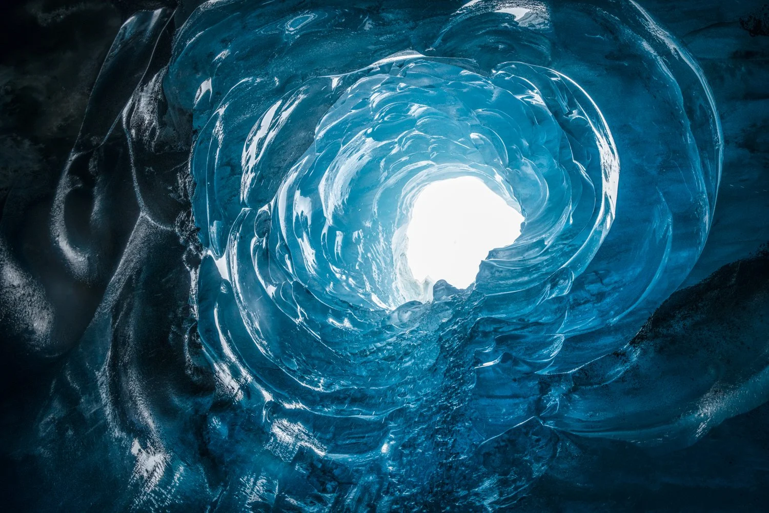 Inside view of an ice cave with a bright opening at the end, walls made of translucent blue ice with curves and textures.