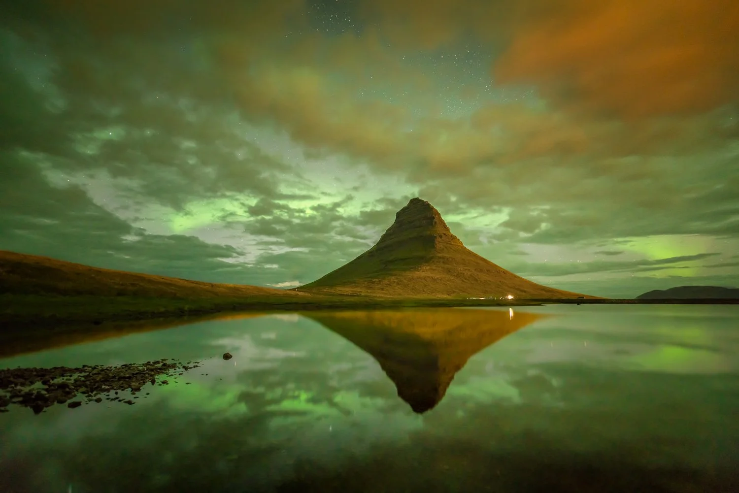 Nighttime view of Kirkjufell mountain reflecting in a calm body of water, with green aurora lights and a cloudy sky overhead.