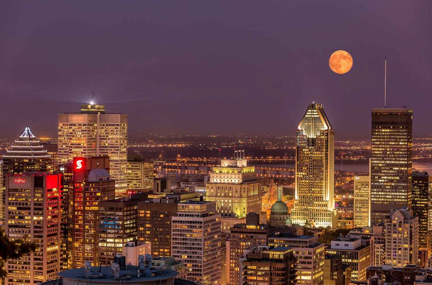 City skyline at dusk with illuminated skyscrapers, a bright half moon in the sky, and purple evening sky backdrop.