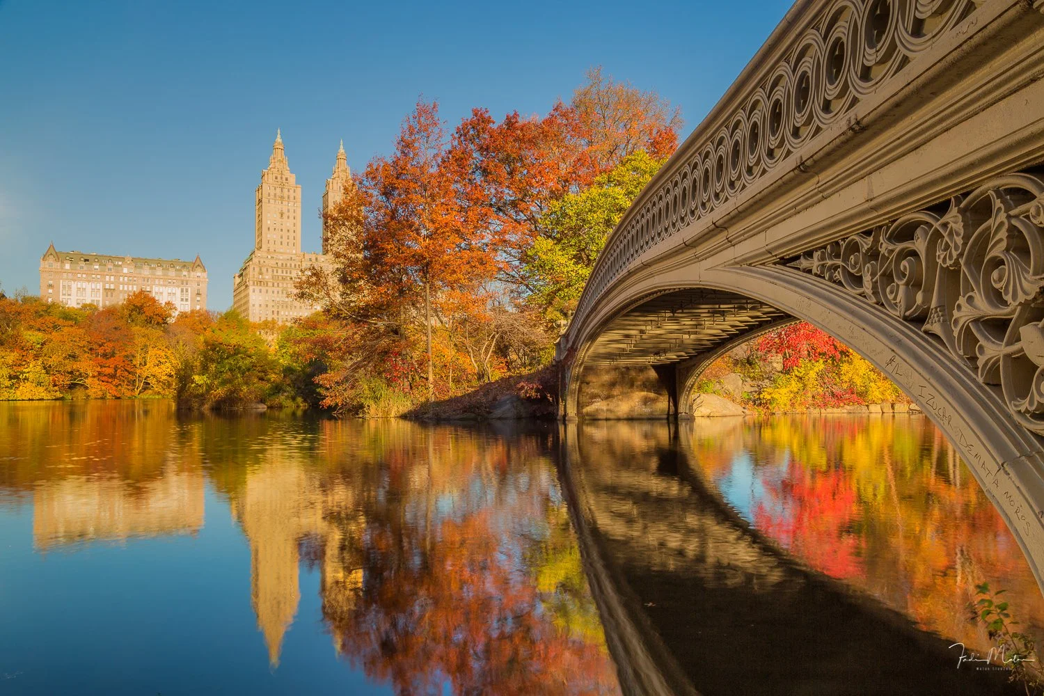 A scenic view of Bow Bridge in Central Park with fall foliage, a decorative bridge over a lake, and tall buildings in the background under a clear blue sky.