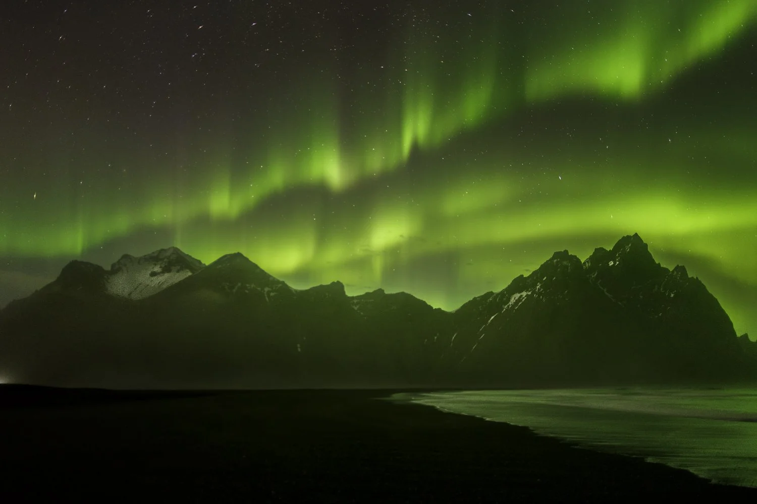 Northern lights glowing green over Vestrahorn mountainous landscape at night with stars in the sky.