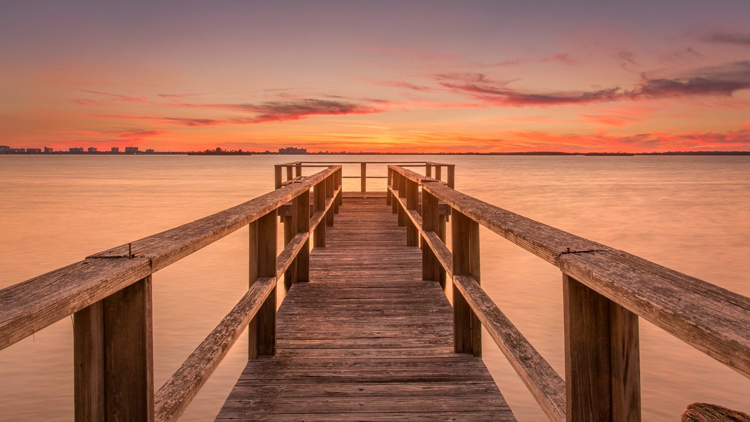 A wooden dock extending over calm water of Dunedin, FL during a colorful sunset with a distant city skyline, Clearwater Beach.