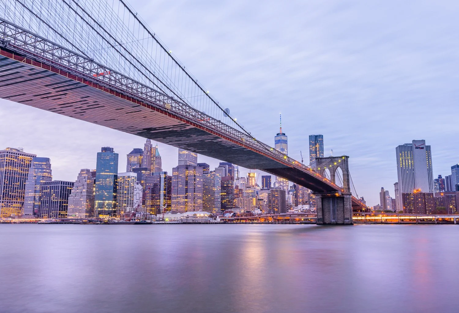 Long exposure photo of the Brooklyn Bridge with New York City skyline in the background during twilight, with some lights reflected in the water.