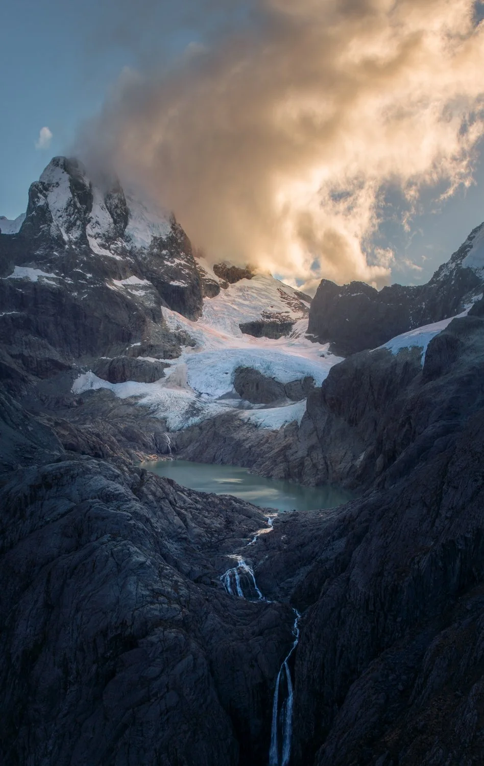 Snow-covered mountain with glaciers, a small lake, and flowing waterfalls surrounded by dark rocky terrain, with a partly cloudy sky and sunlight. Shot taken by areal photography in New Zealand