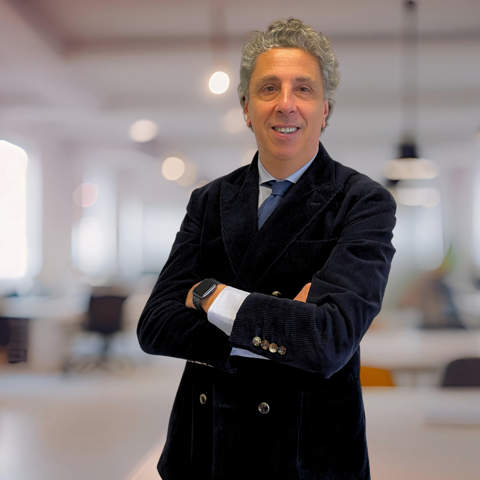 A smiling man with gray curly hair wearing a dark blazer, white shirt, and blue tie standing with arms crossed in a bright, modern office space. Pierluca Lombardi