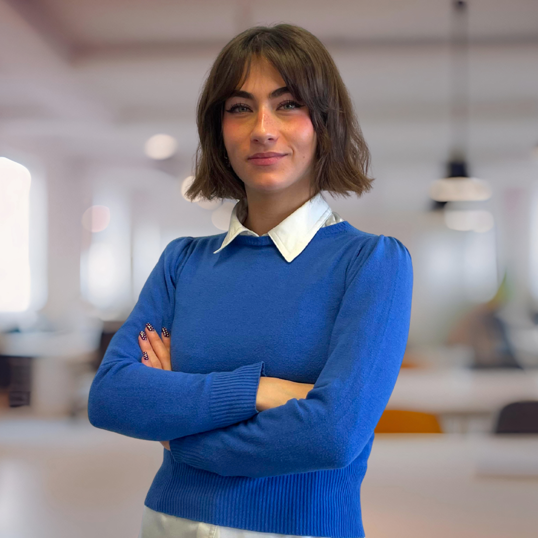 A confident woman with short brown hair is standing with her arms crossed in a modern office space, wearing a blue sweater over a white collared shirt. Chiara Greco