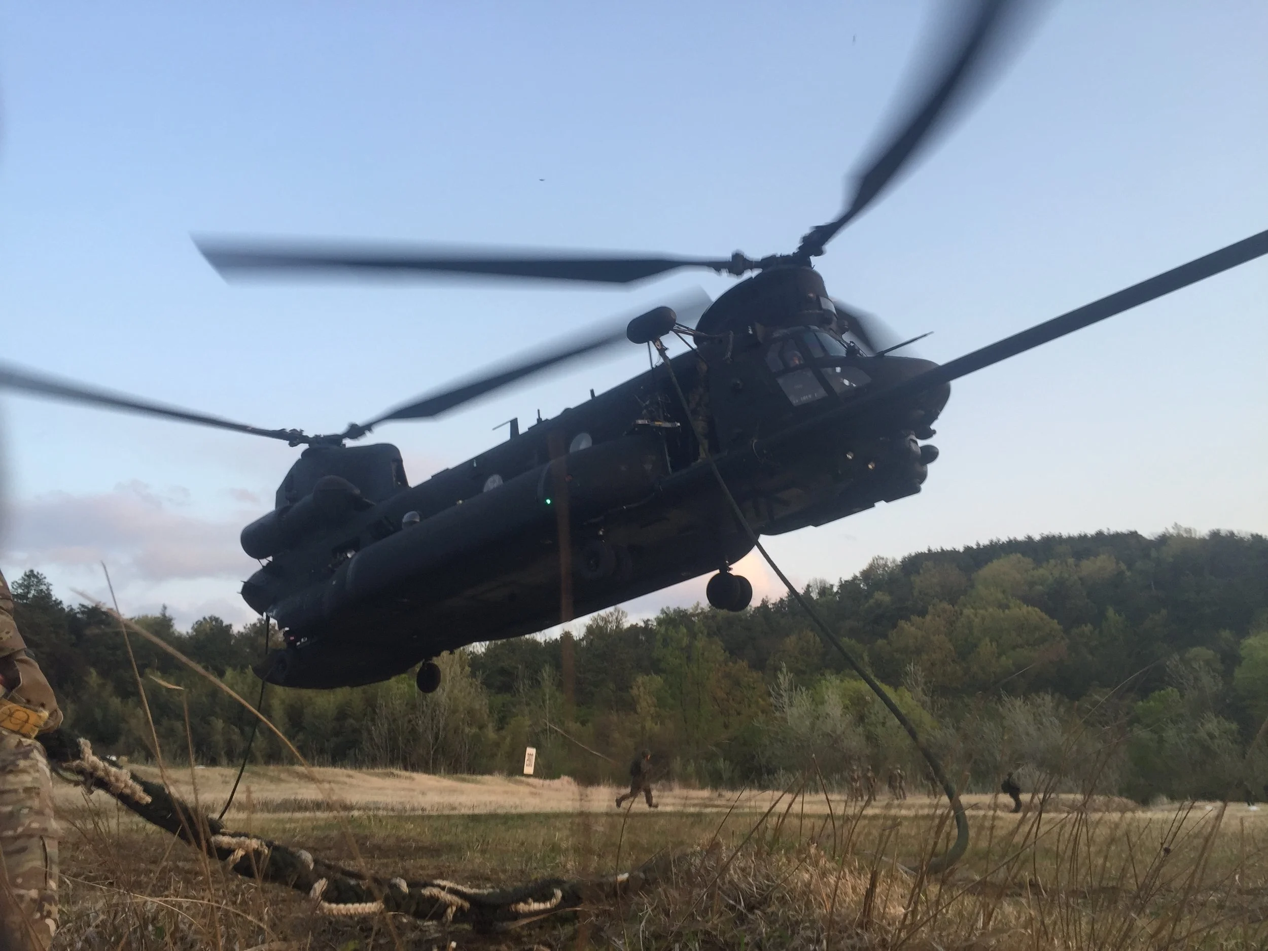 Black military helicopter flying low over a field with trees in the background, with soldiers visible in the distance and grassy terrain in the foreground.