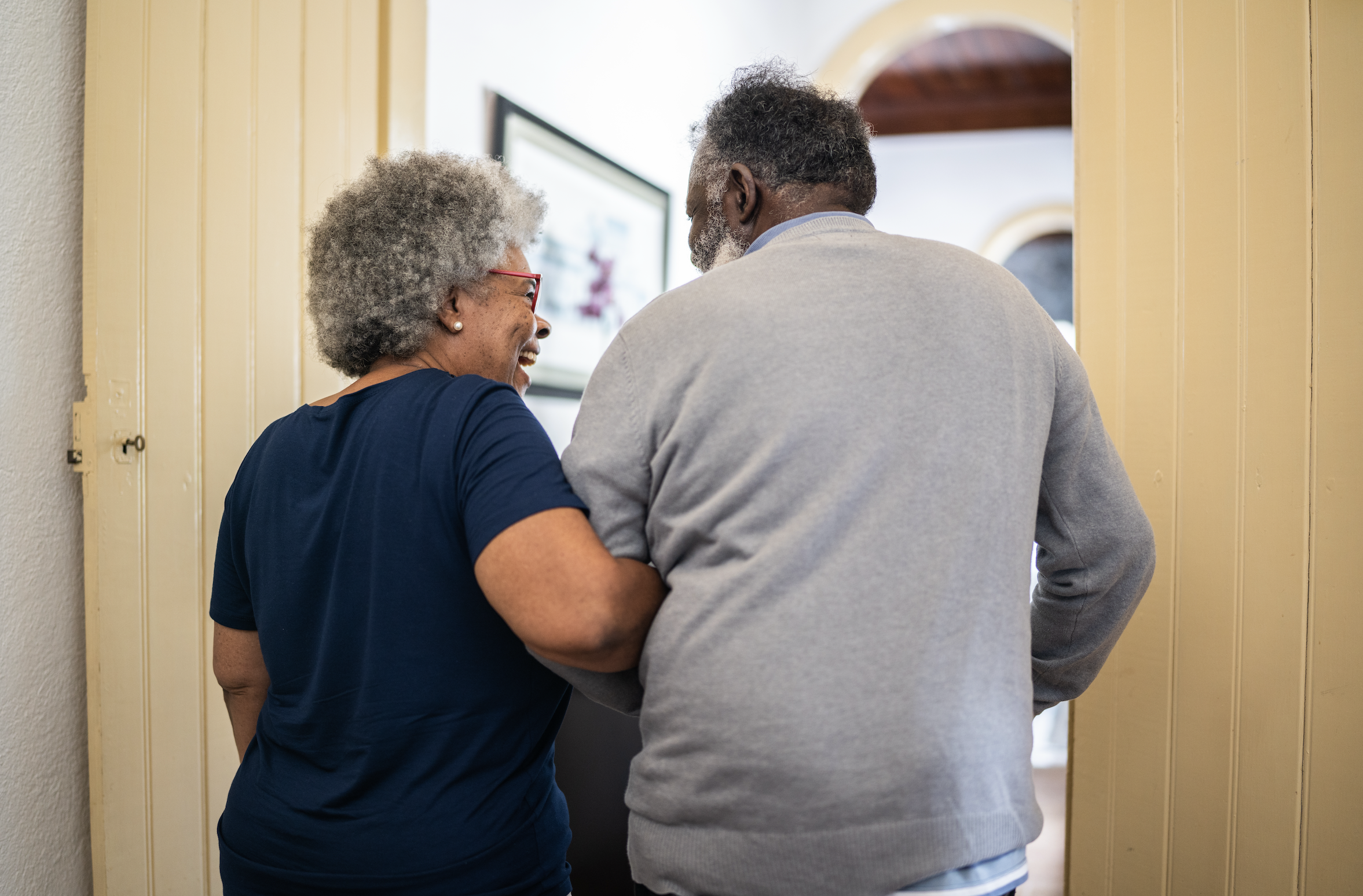 An elderly woman and man are sharing a happy moment, standing close together. The woman is smiling and wearing a navy blue shirt, glasses, and earrings, while the man, with gray hair and beard, is wearing a light gray sweater. They are standing in a hallway near an open door, with picture frames on the wall behind them.